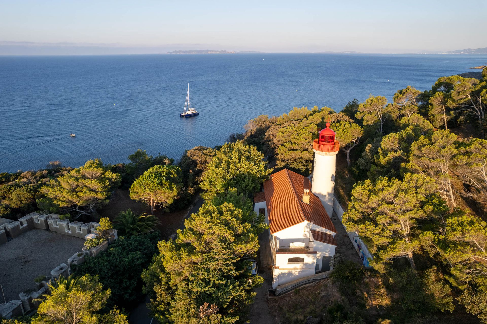Catamaran offshore near a wooded Mediterranean headland and lighthouse