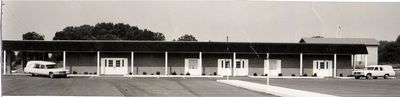 Black-and-white photo of a long roadside building with a central entrance, parked cars, and trees behind it.