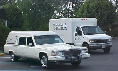 White hearse and box truck with “Mannsville Funeral Home” parked on a road with trees in the background