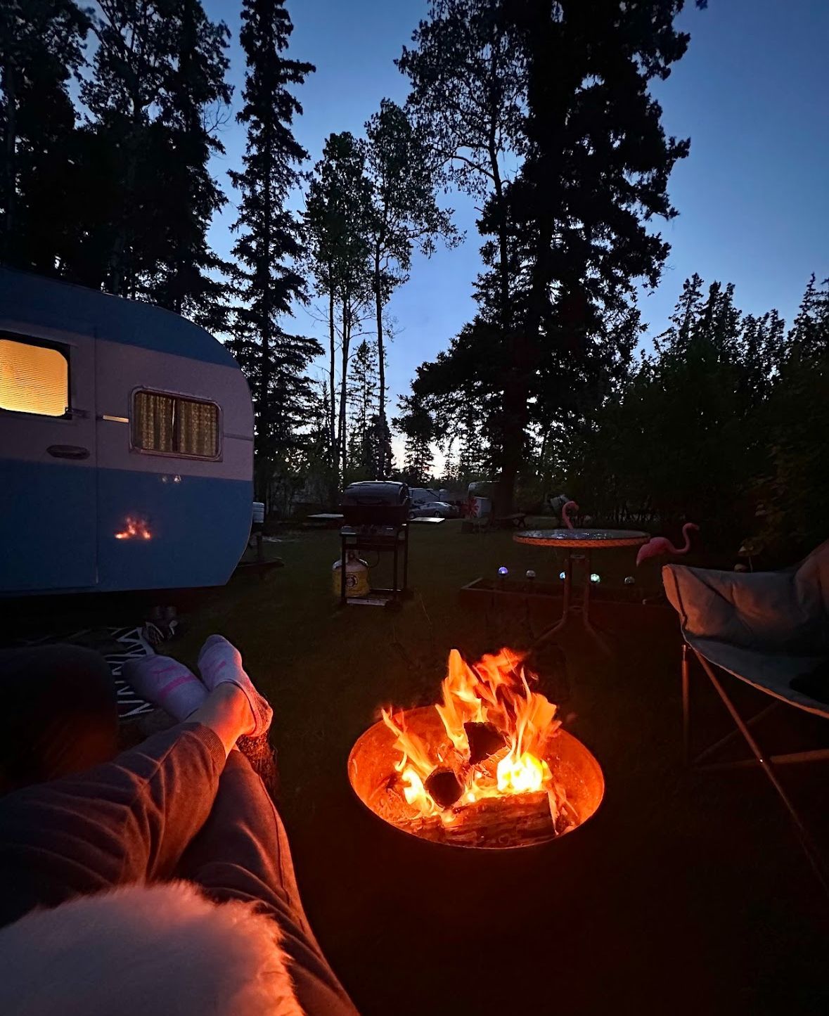 A person sitting in front of a campfire with a trailer in the background