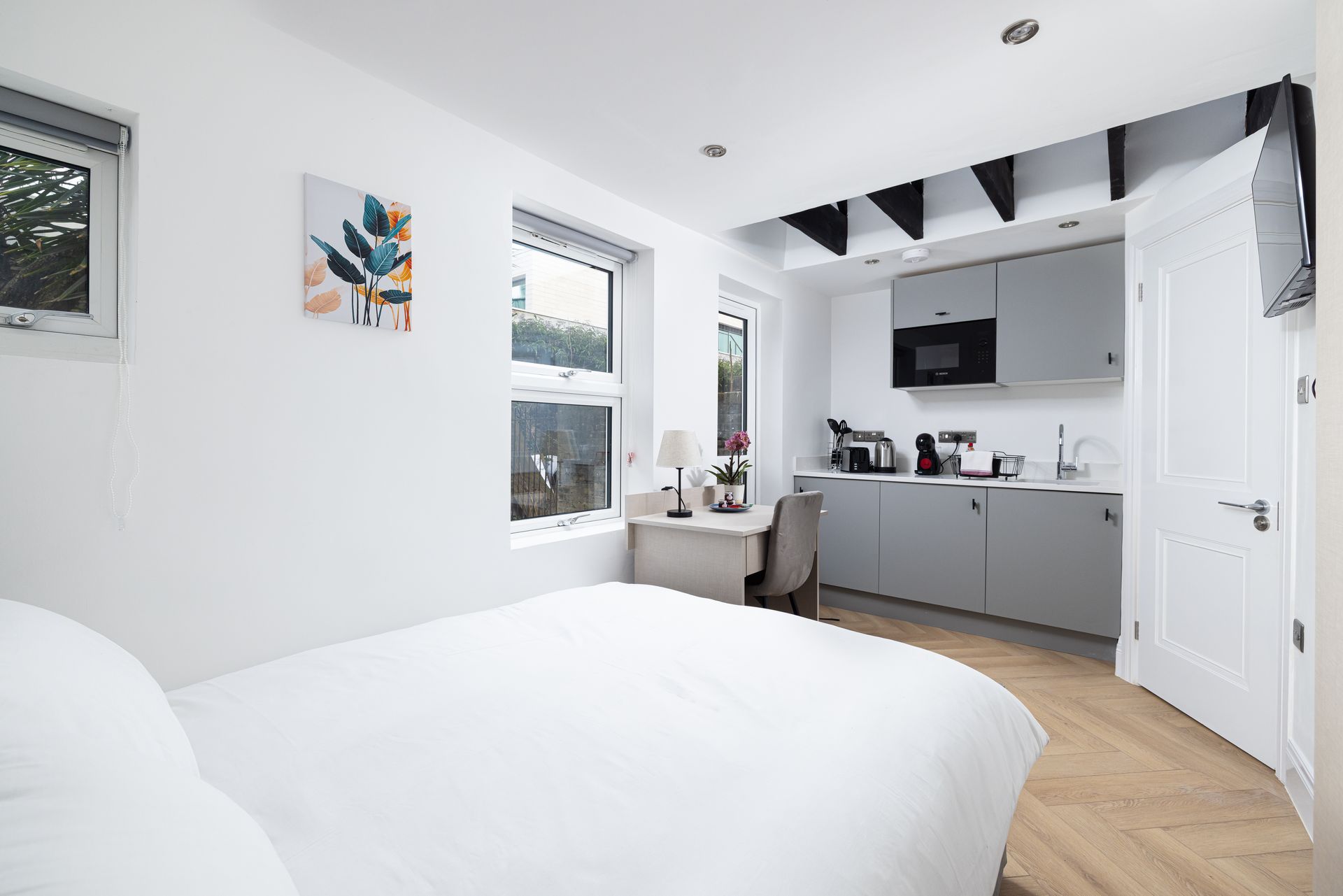 Bright white bedroom with a bed, windows, and a small open kitchen area beyond.