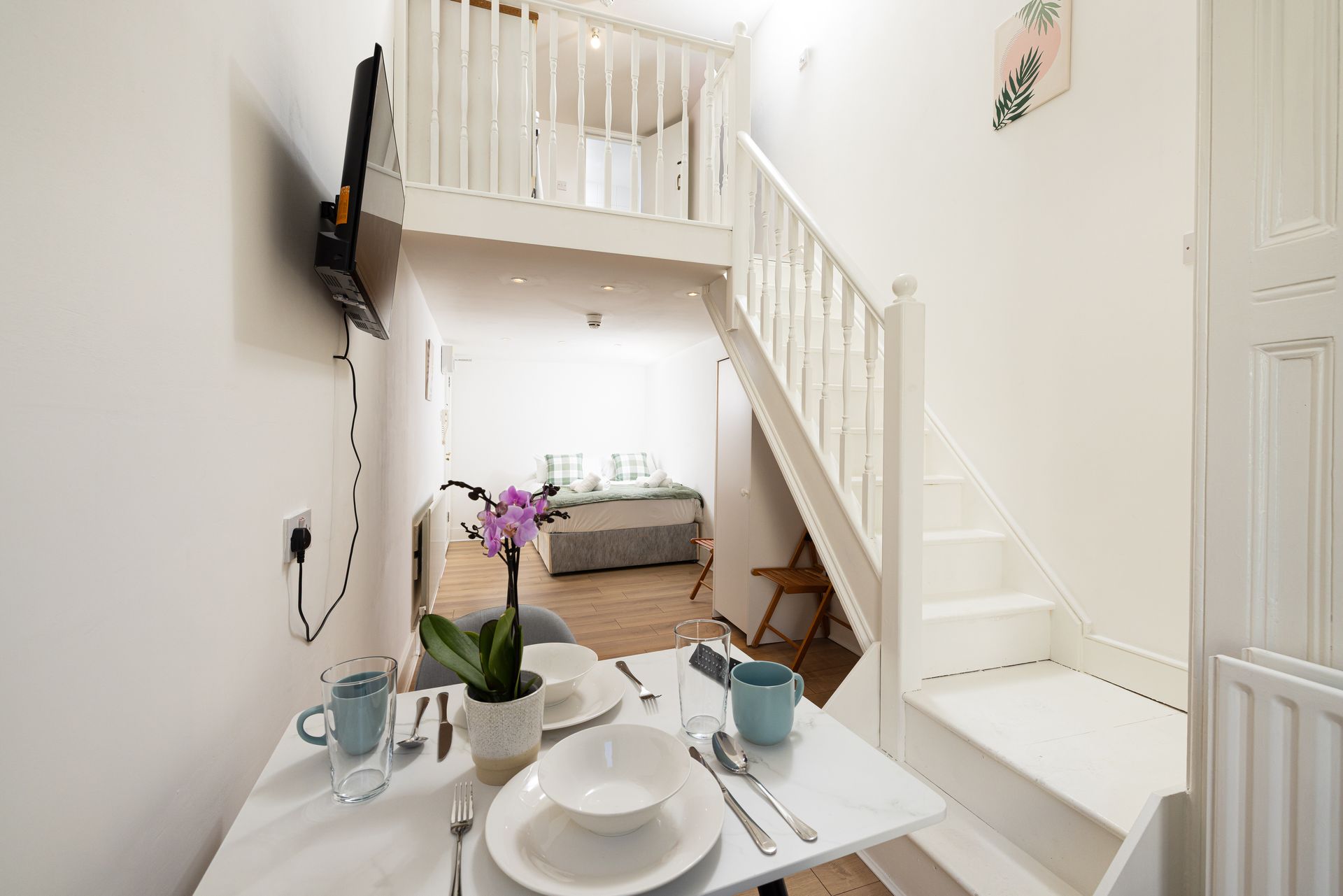 Bright white dining nook with a set table, staircase, wall-mounted TV, and cozy sitting area beneath a loft.