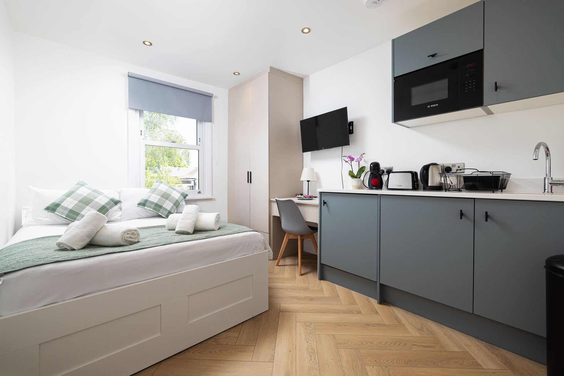 Compact modern studio bedroom with gray kitchenette, bed, desk, and large window with blinds