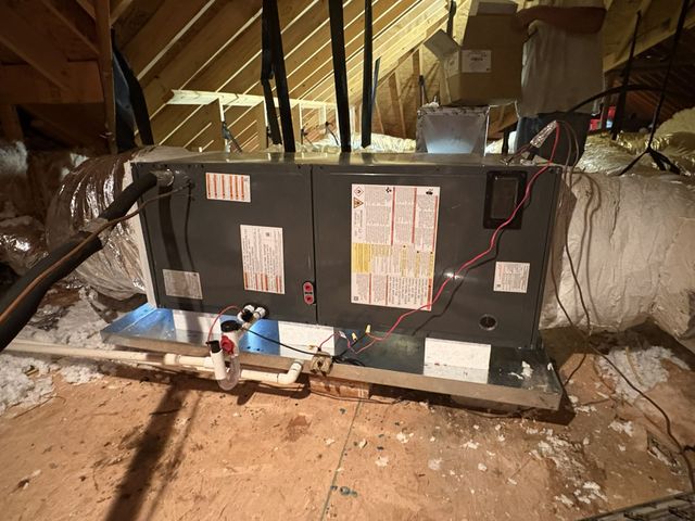 Black HVAC unit in an attic with exposed insulation, pipes, and wiring on a wooden floor.