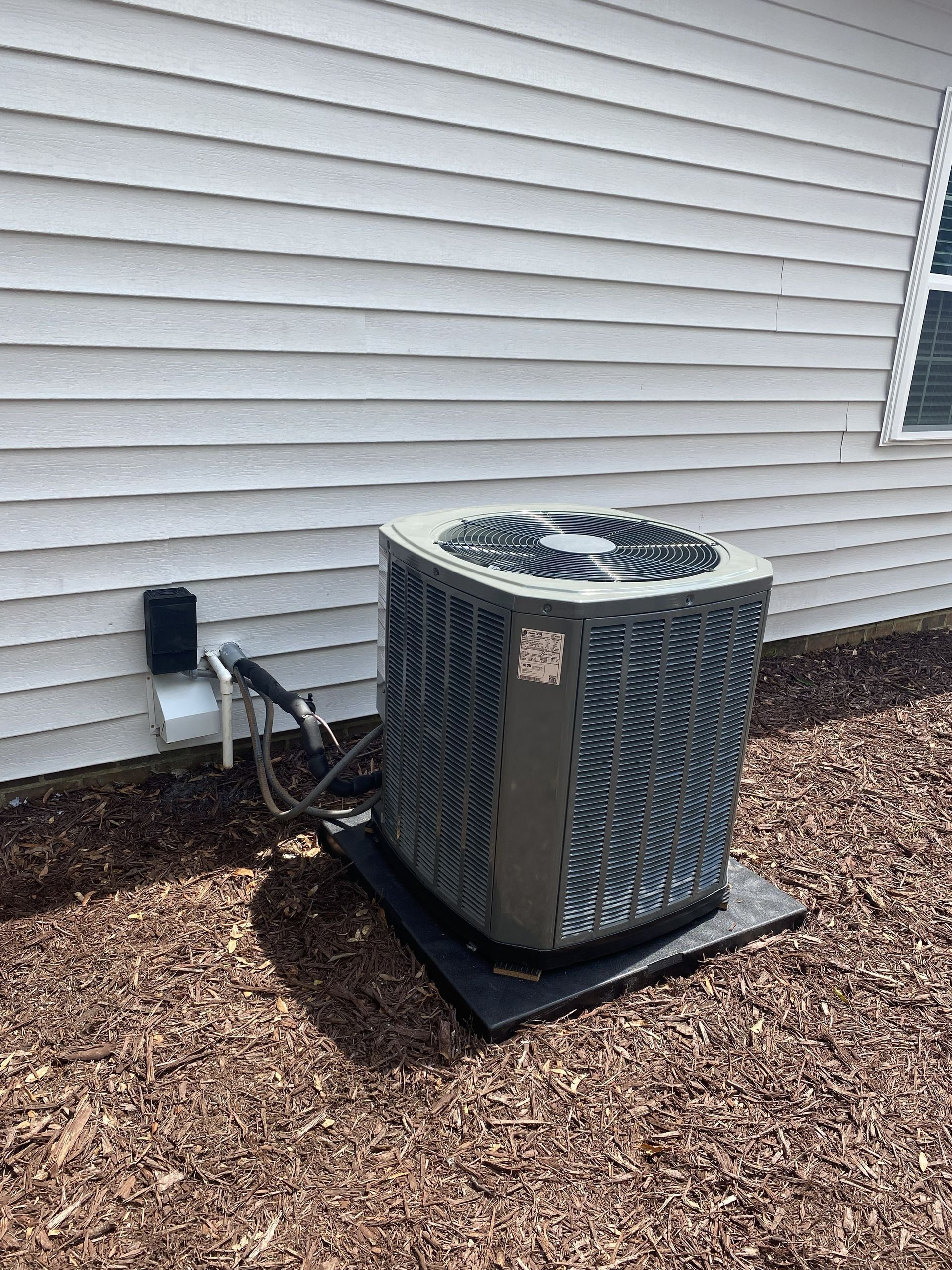 Outdoor air conditioner unit beside a house with white siding and mulch bed