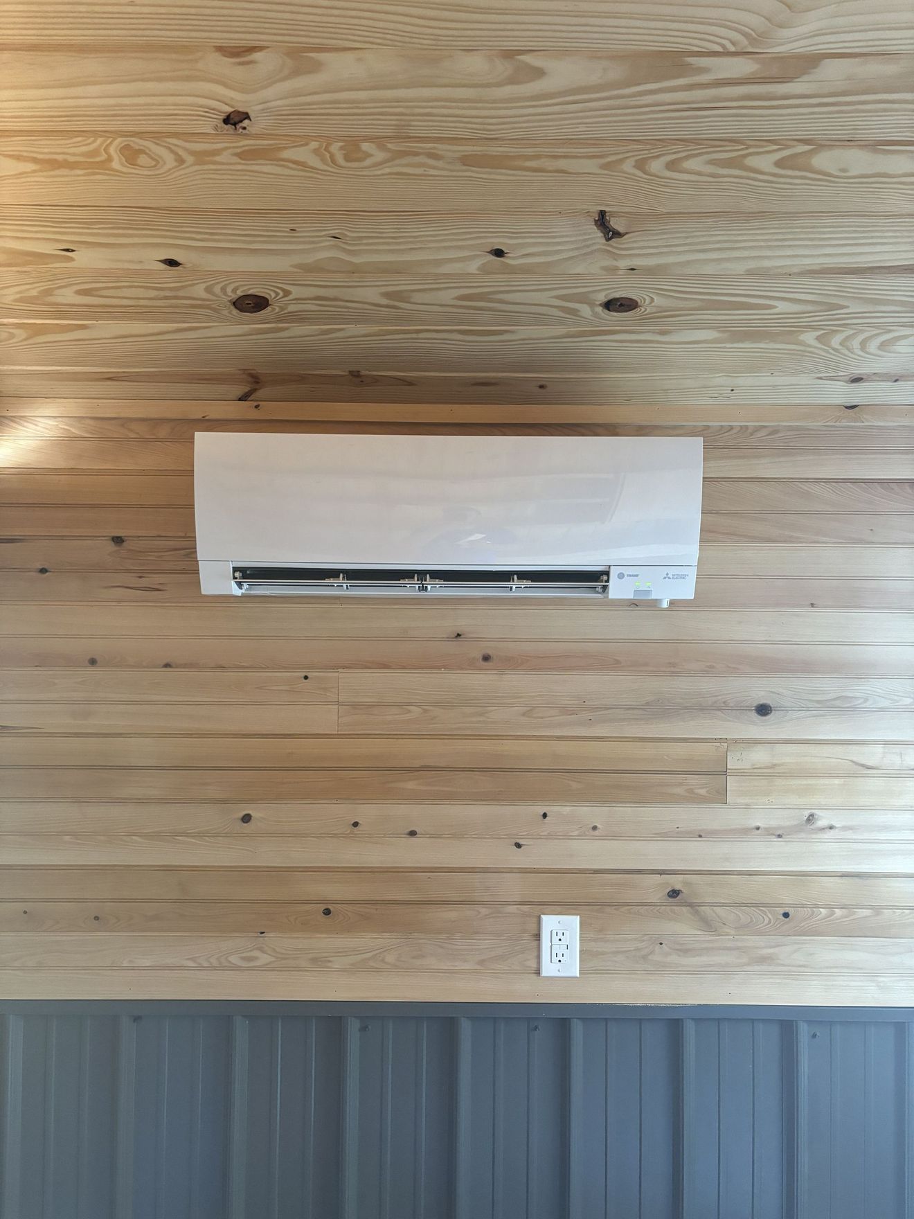 Wall-mounted white air conditioner on a wood-paneled wall with gray wainscoting and an outlet below