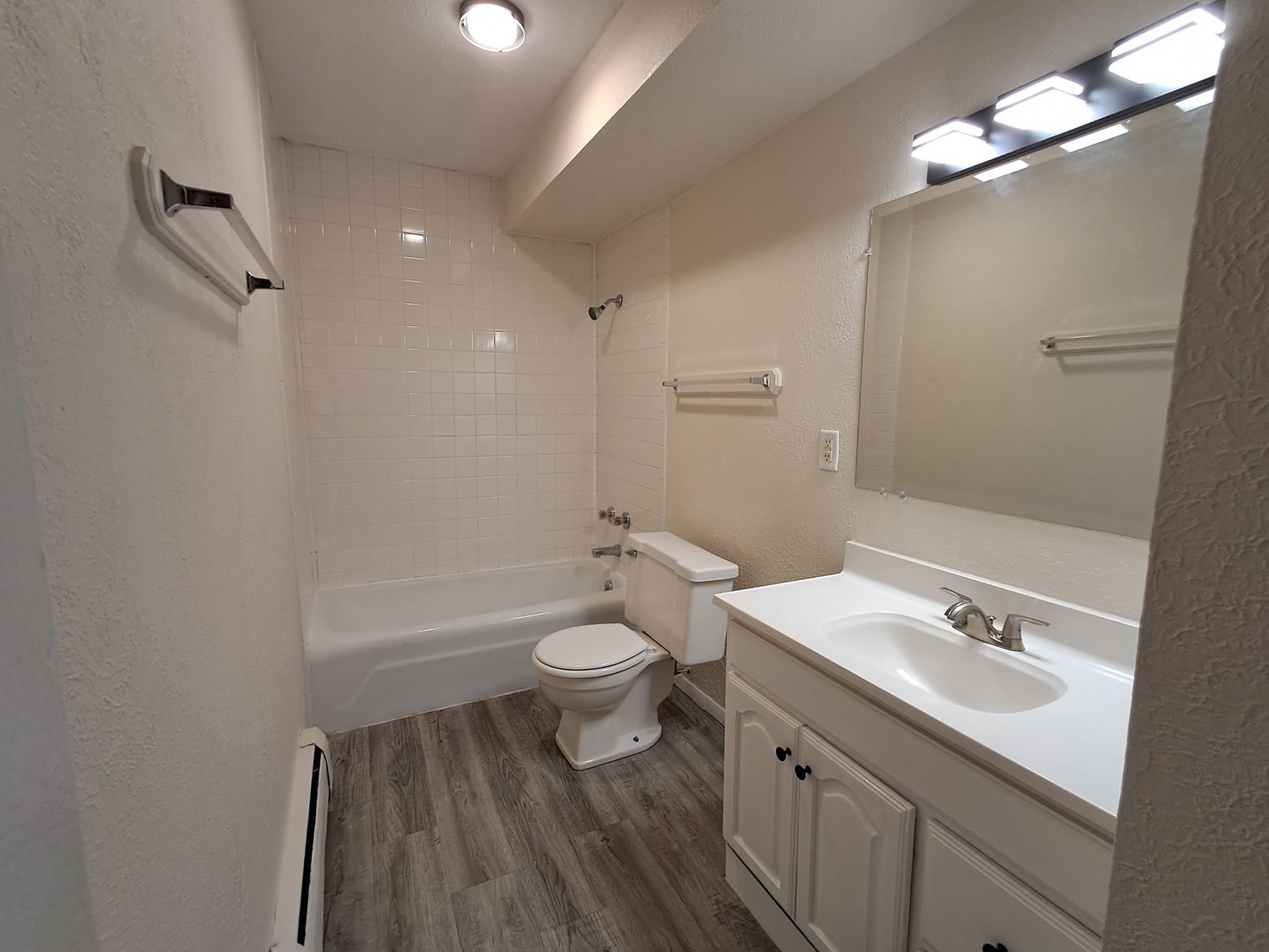 Small white bathroom with tub, toilet, vanity, mirror, and wood-look floorboards.