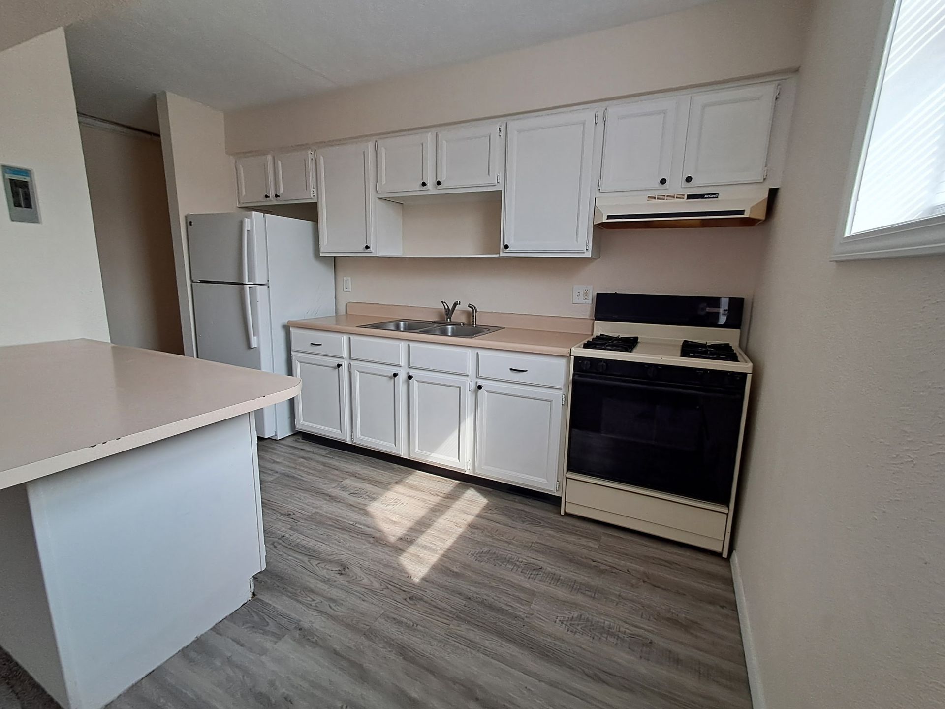 Empty kitchen with white cabinets, refrigerator, stove, and beige countertops in soft sunlight