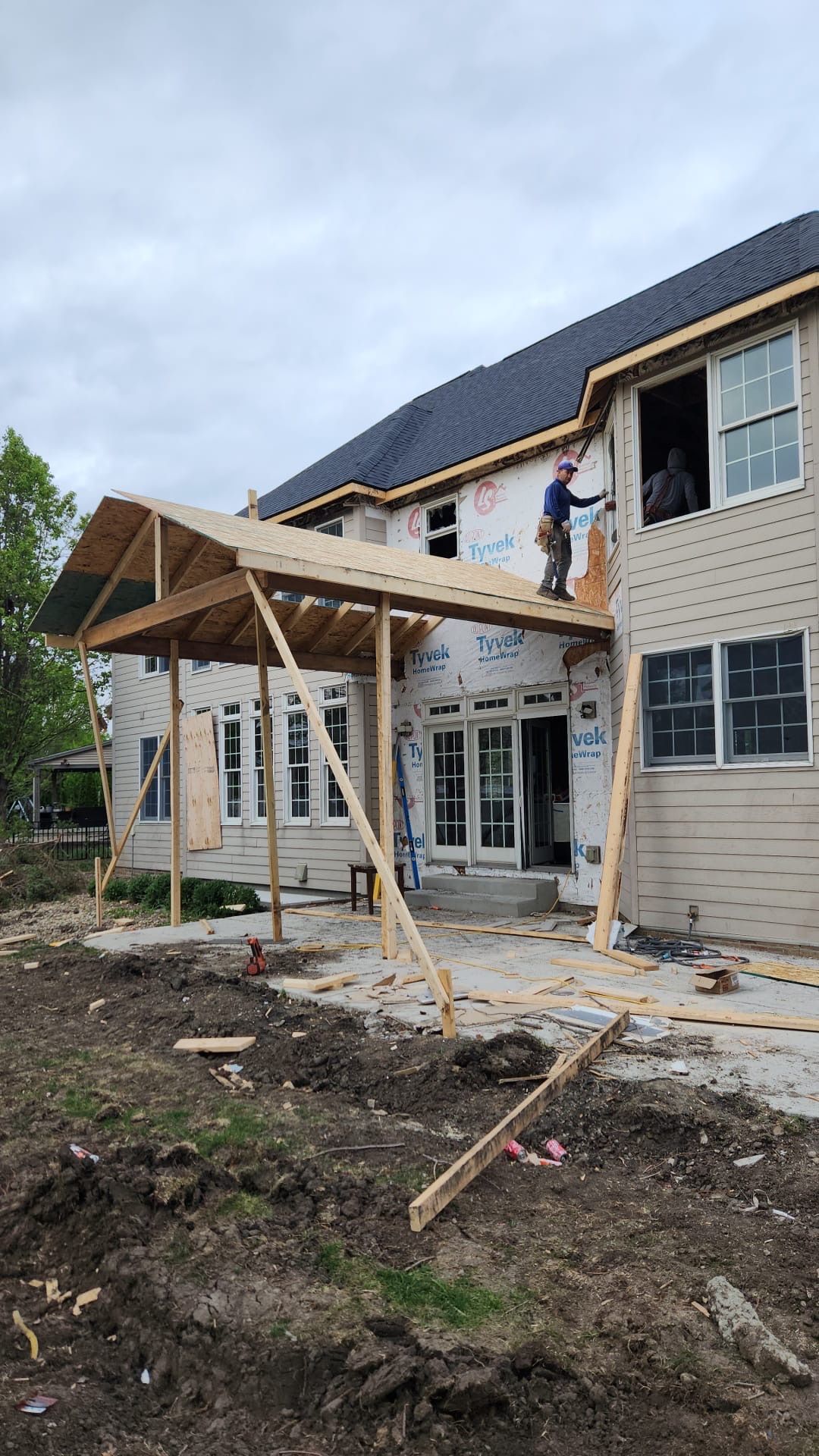 Two workers build a wooden porch roof on the side of a house under construction.