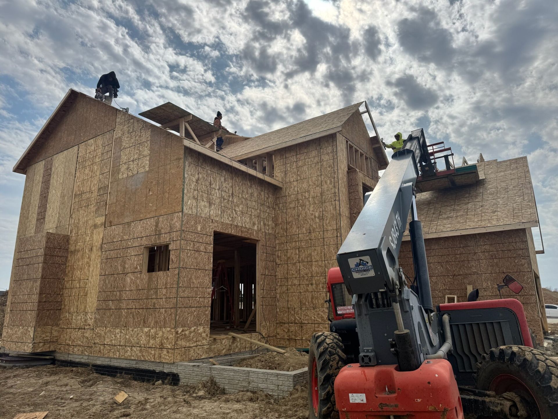 Workers and a red lift truck on a house under construction with plywood sheathing and roof framing