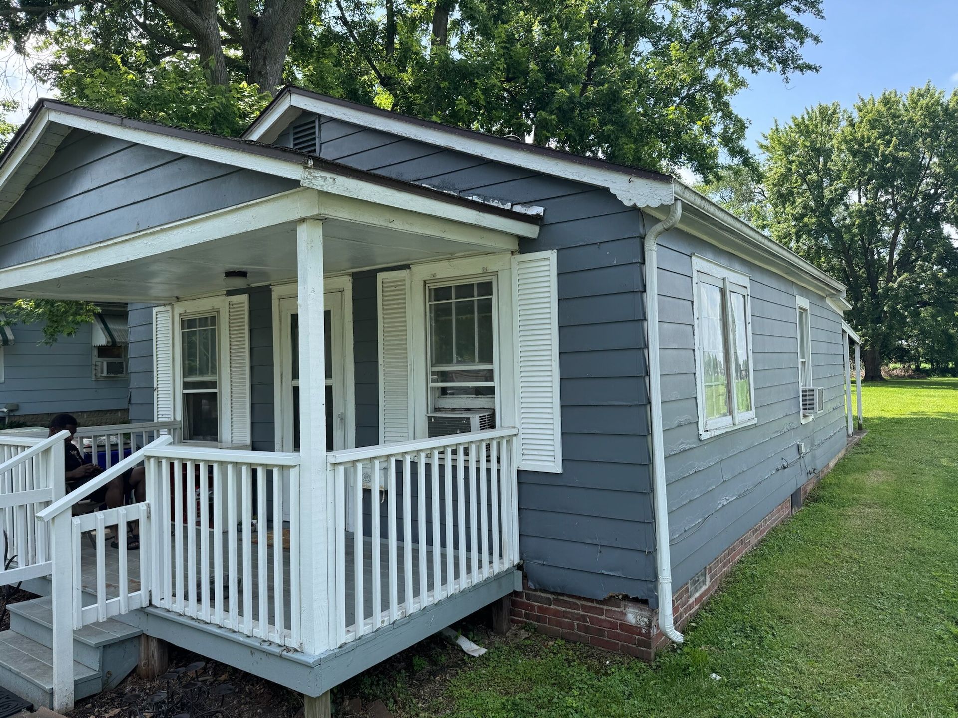 Small gray house with white porch railing and steps, surrounded by trees and grass.