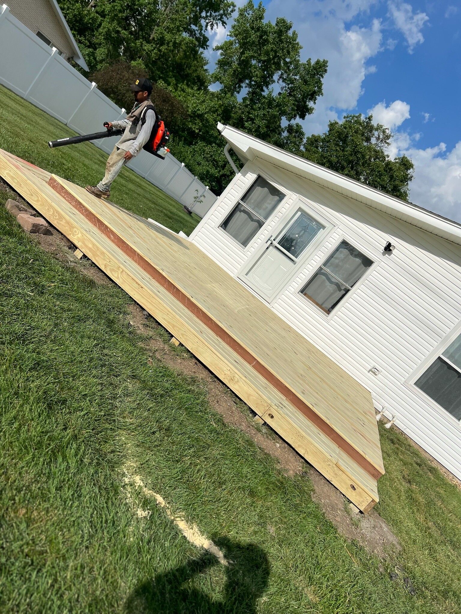 Worker leveling wooden boards beside a white house on a grassy yard under a blue sky.