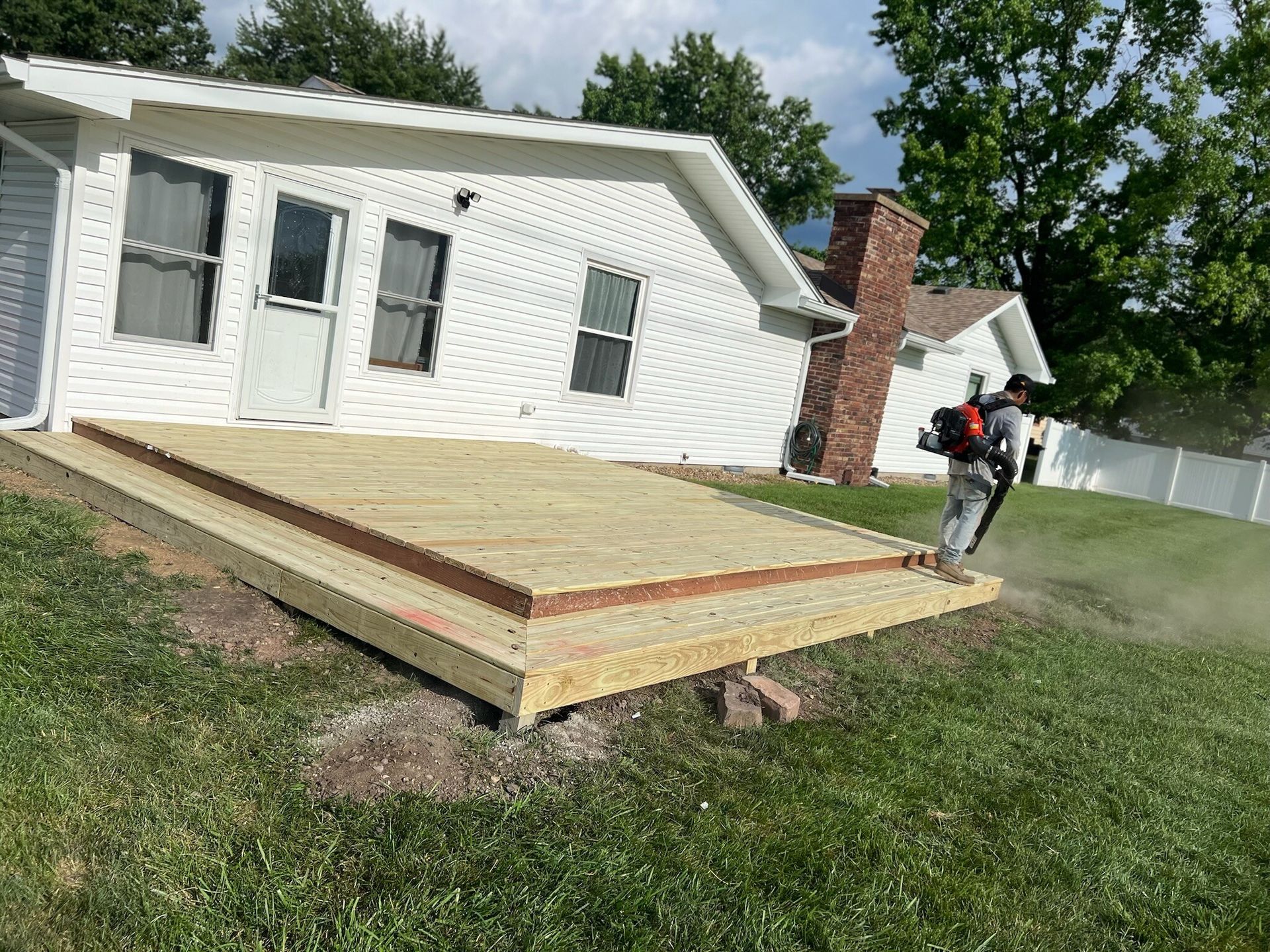 New front porch deck under construction beside a white house, with one worker using a blower.