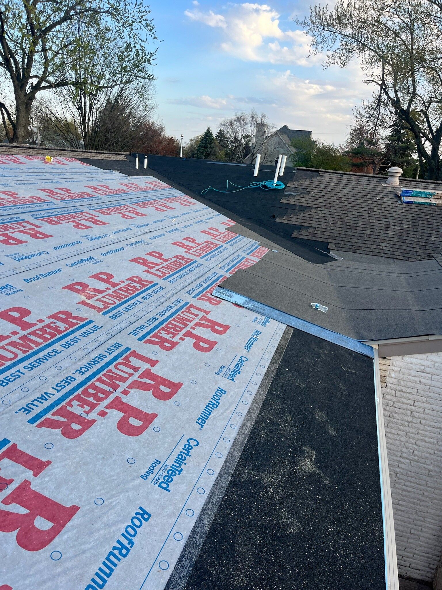 Roofer installing black shingles beside white underlayment on a pitched roof under a blue sky