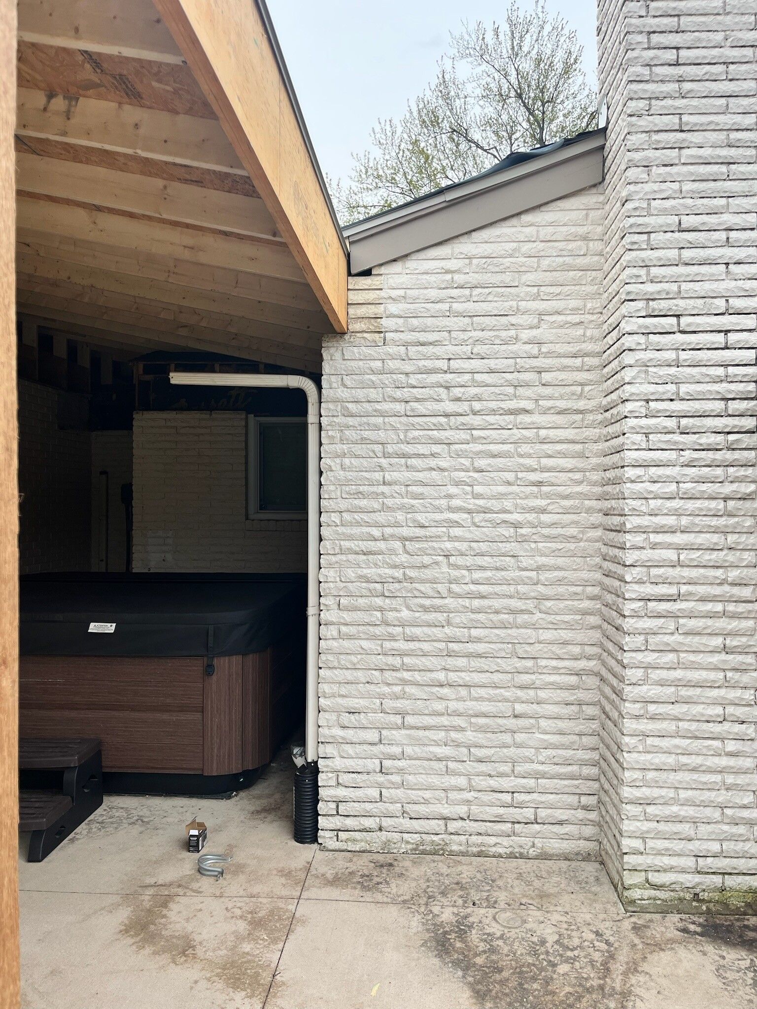 Covered patio beside a white brick house, with a hot tub under the roof and a small dog on the concrete.