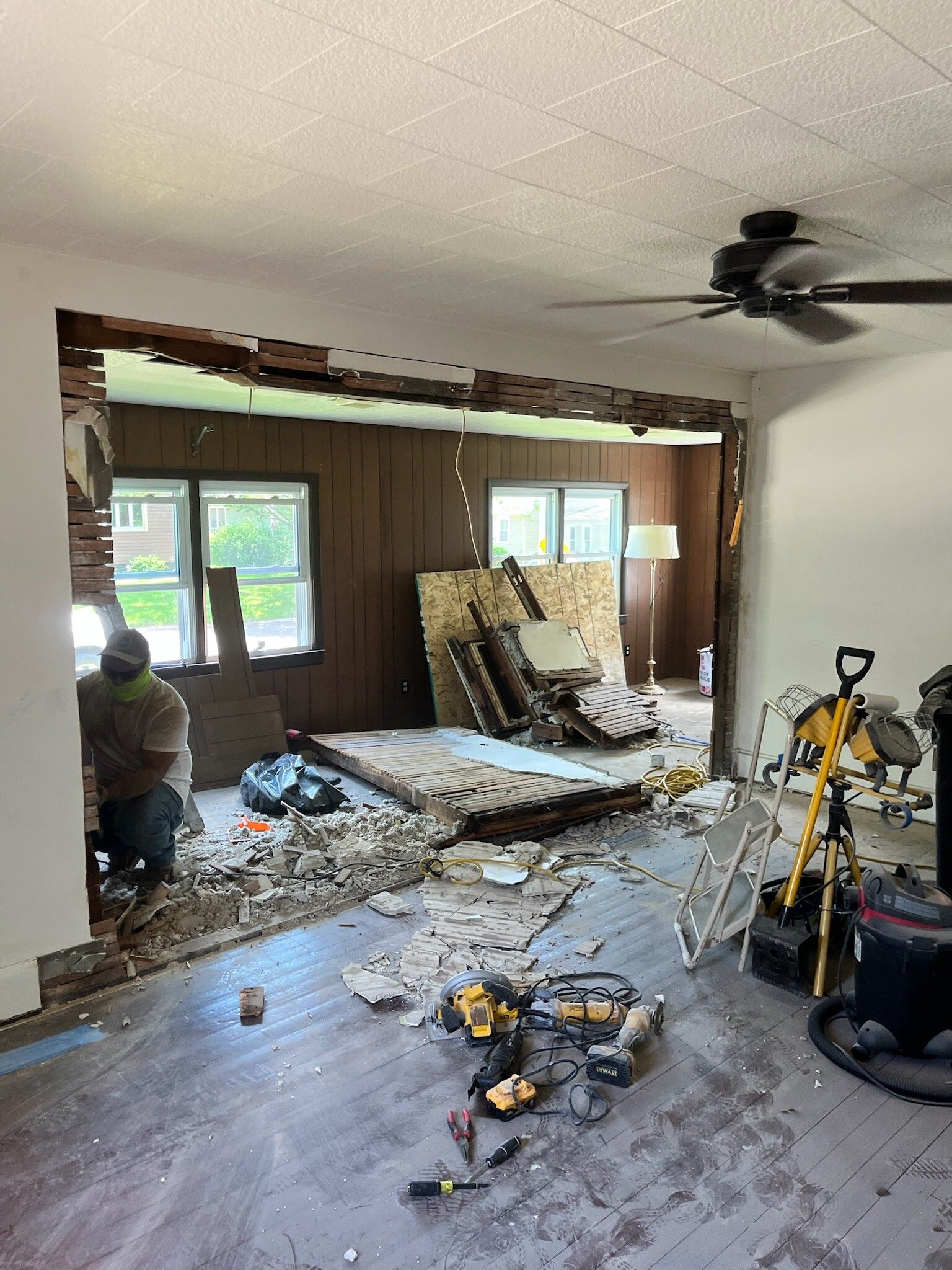 Damaged living room under renovation, with rubble, exposed wall studs, and tools scattered on the floor.