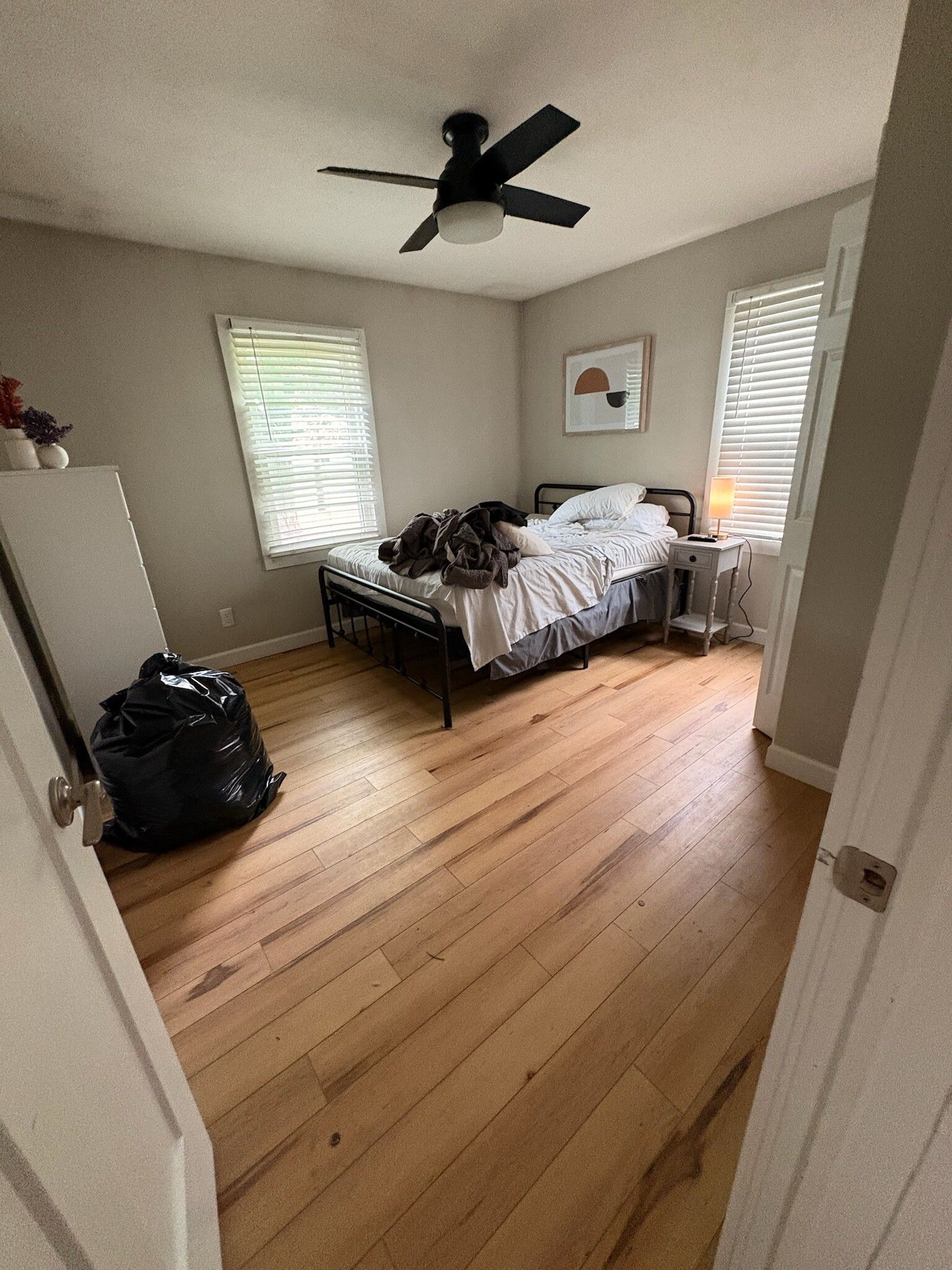 Bedroom with wooden floors, black ceiling fan, white walls, two windows, and an unmade bed near the back wall