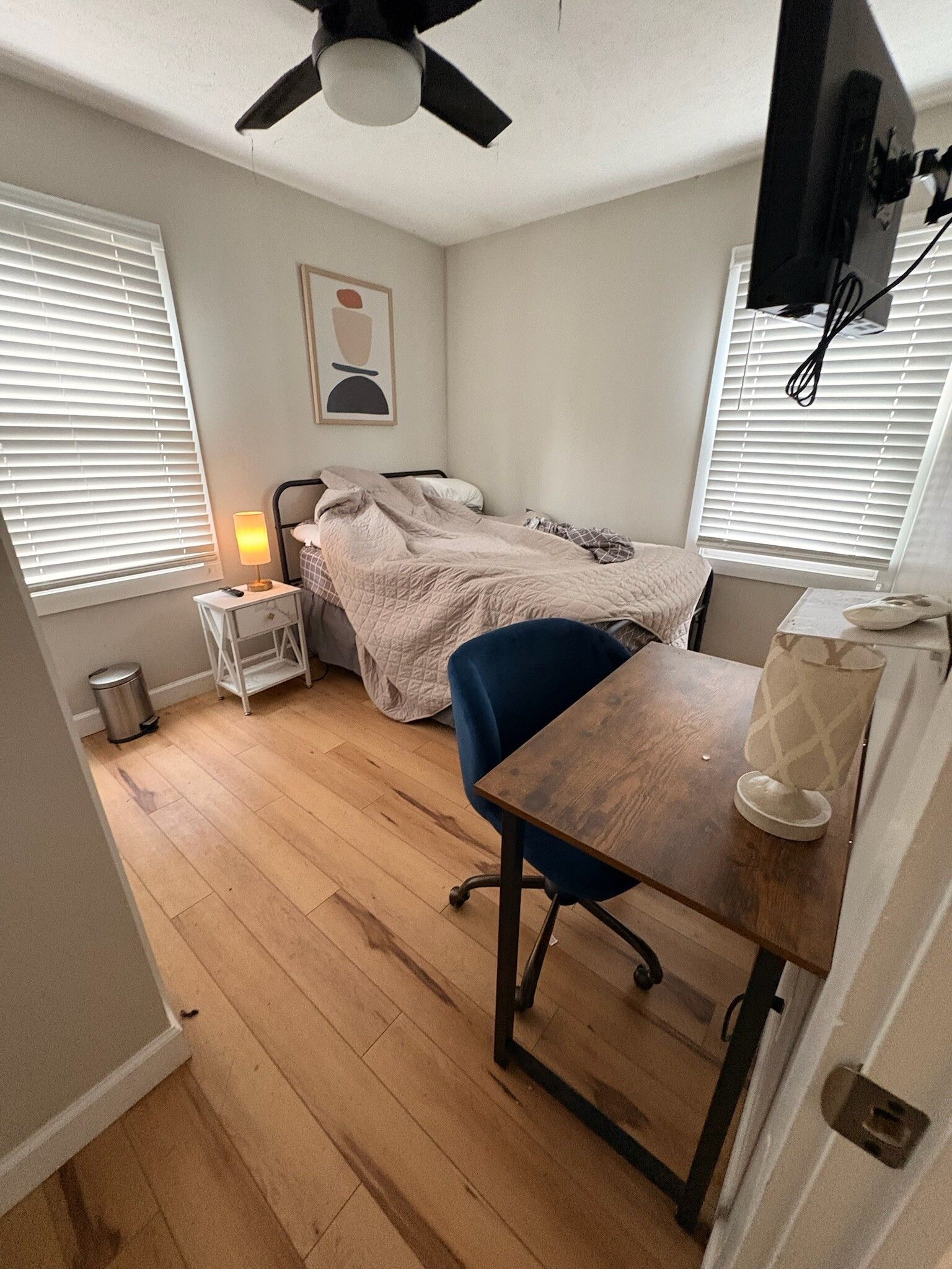 Small bedroom with an unmade bed, desk chair, nightstand lamp, wall art, and windows with blinds.