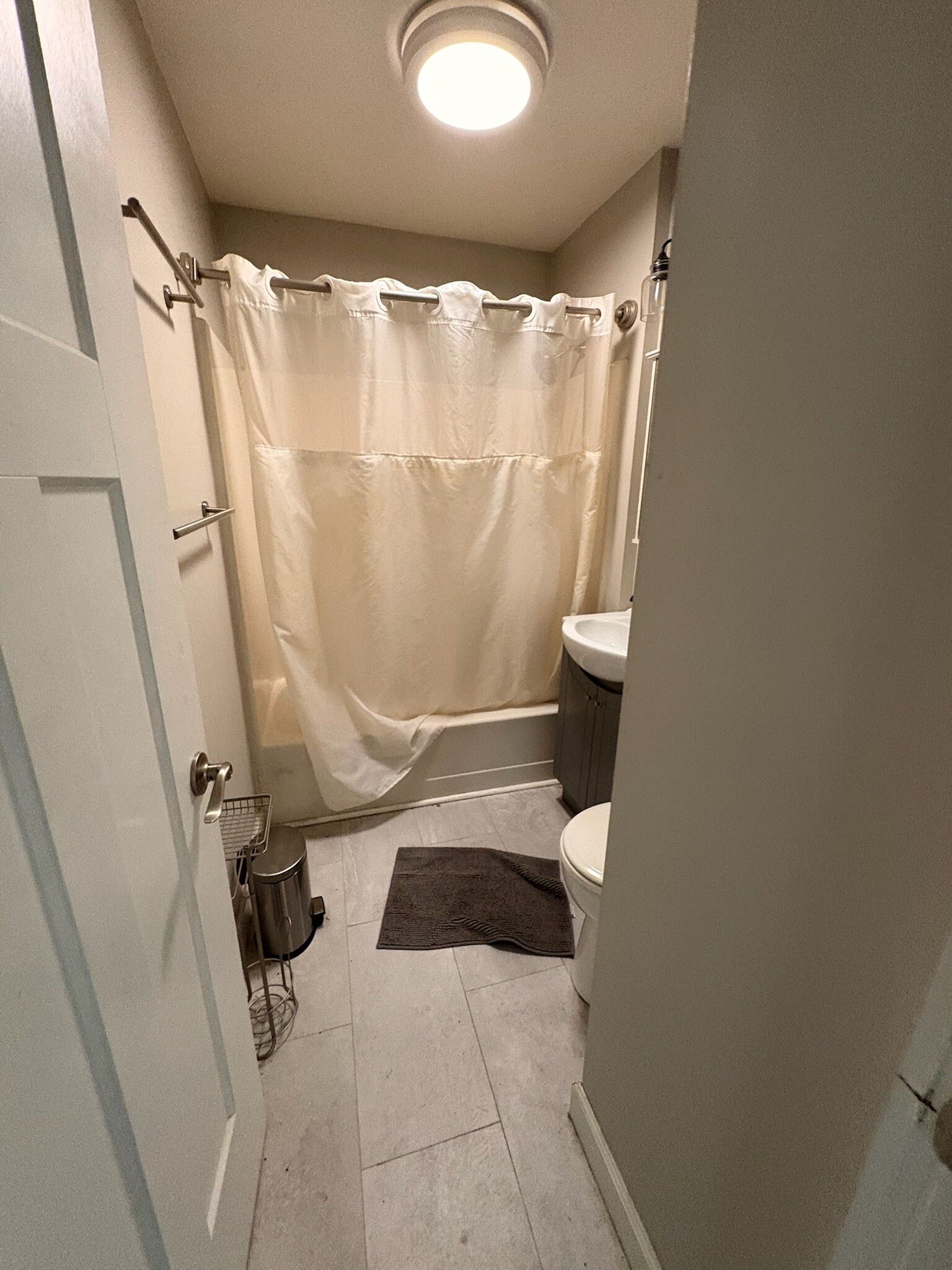 Narrow bathroom with beige shower curtain, gray floor rug, vanity sink, and ceiling light.