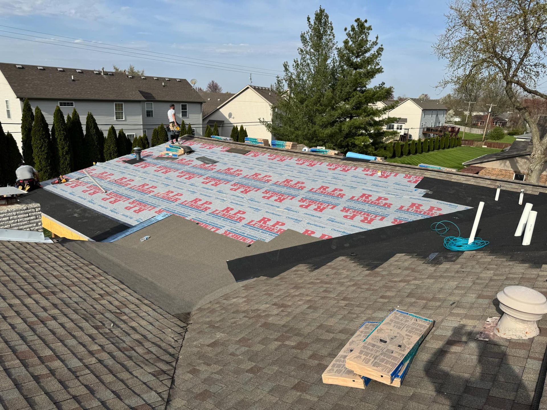 Roof under repair with red and blue underlayment on a suburban house, surrounded by trees and neighboring homes