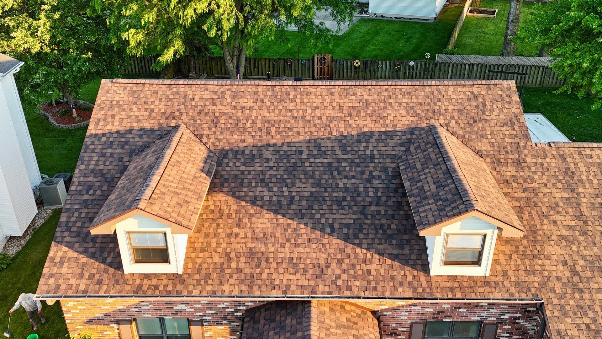 Aerial view of a house roof with brown shingles and two dormer windows, bordered by green trees