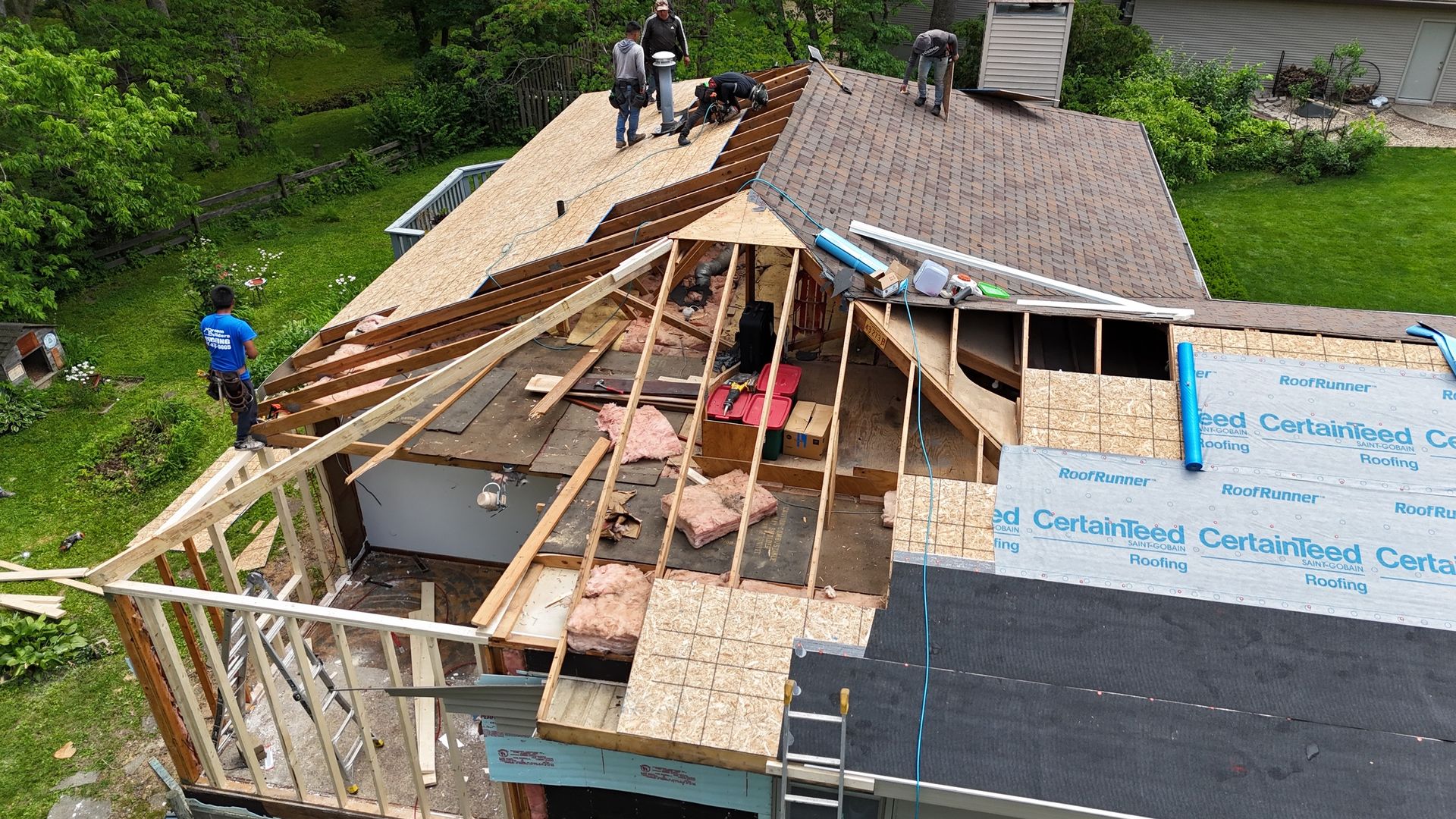 Workers repairing a damaged roof on a house in a wooded area, with exposed beams and blue tarps