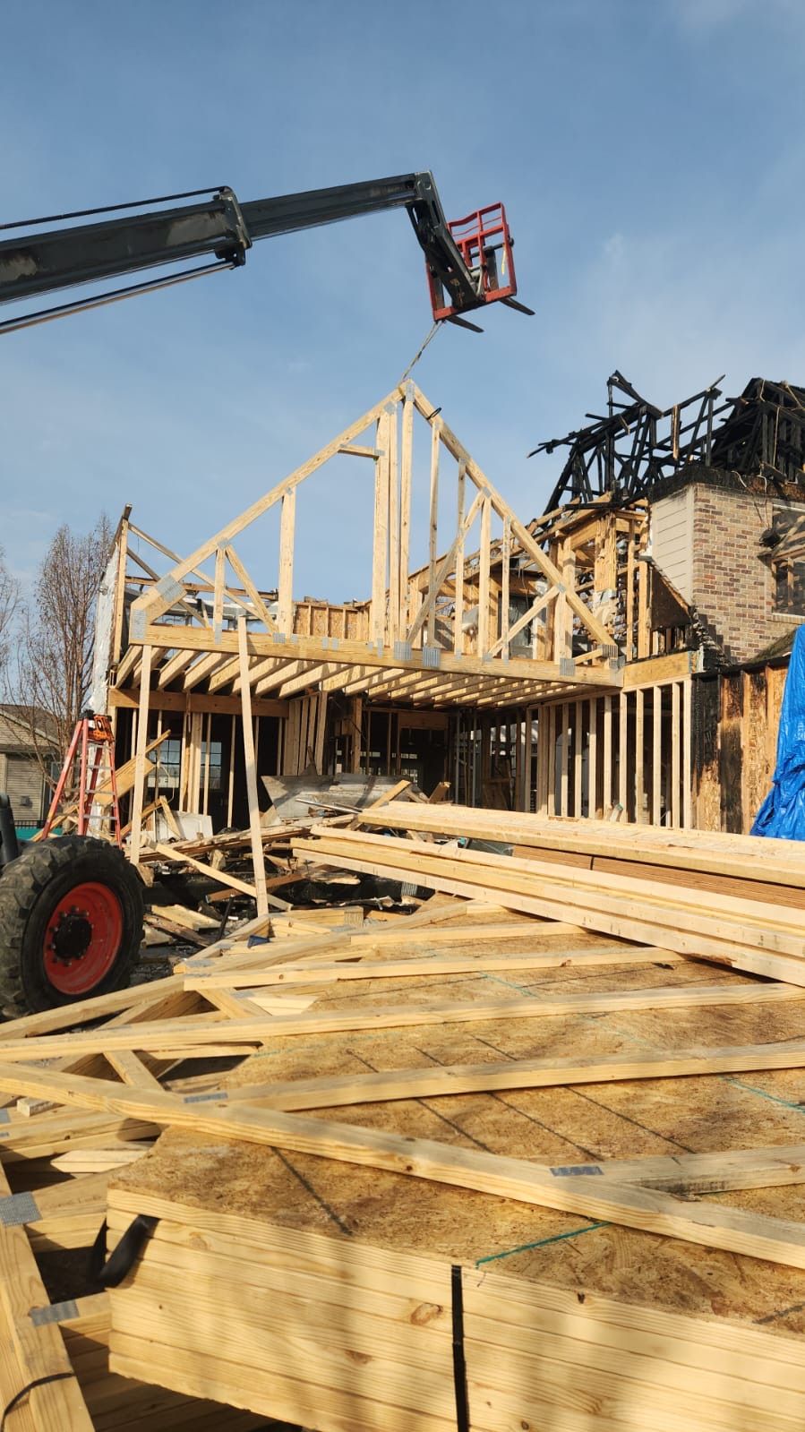 Framing of a house under construction with crane, exposed wood trusses, and lumber stacks in the foreground