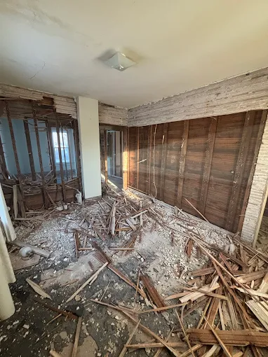 Damaged room with debris scattered across the floor, exposed ceiling, and broken walls and windows