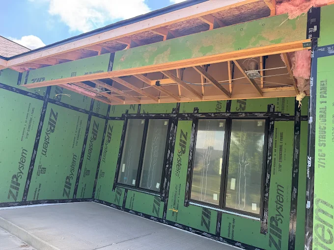 Partially built porch with green sheathing, exposed roof framing, and large windows under a blue sky