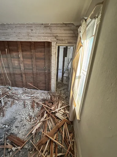 Damaged room with debris on the floor, exposed wall framing, and a broken window letting in daylight