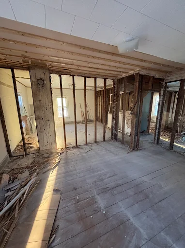 Interior under renovation with exposed wall studs, stripped floorboards, and debris.