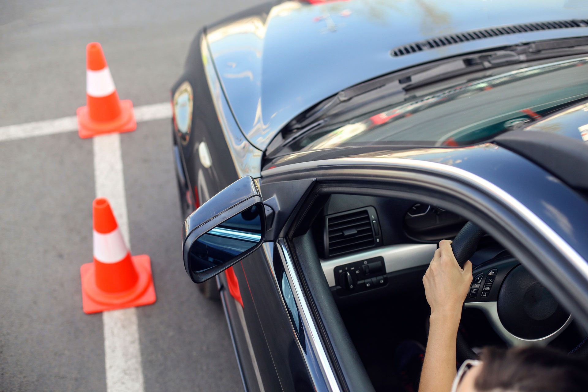 A woman is driving a car in a parking lot next to traffic cones