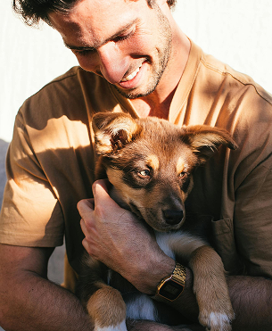 A person in a tan shirt smiles while gently holding a brown and white puppy in their arms.