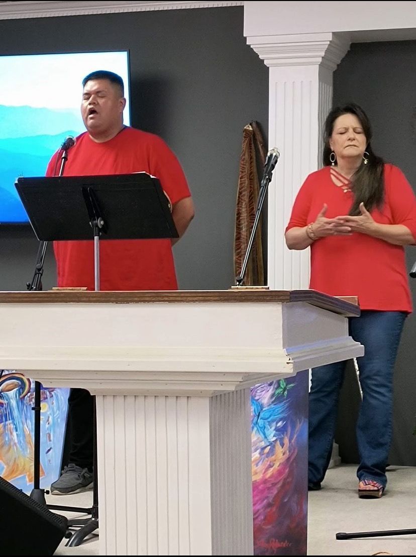 A man in a red shirt is singing into a microphone while a woman stands behind him