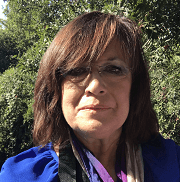A woman wearing glasses and a blue shirt is standing in front of trees.