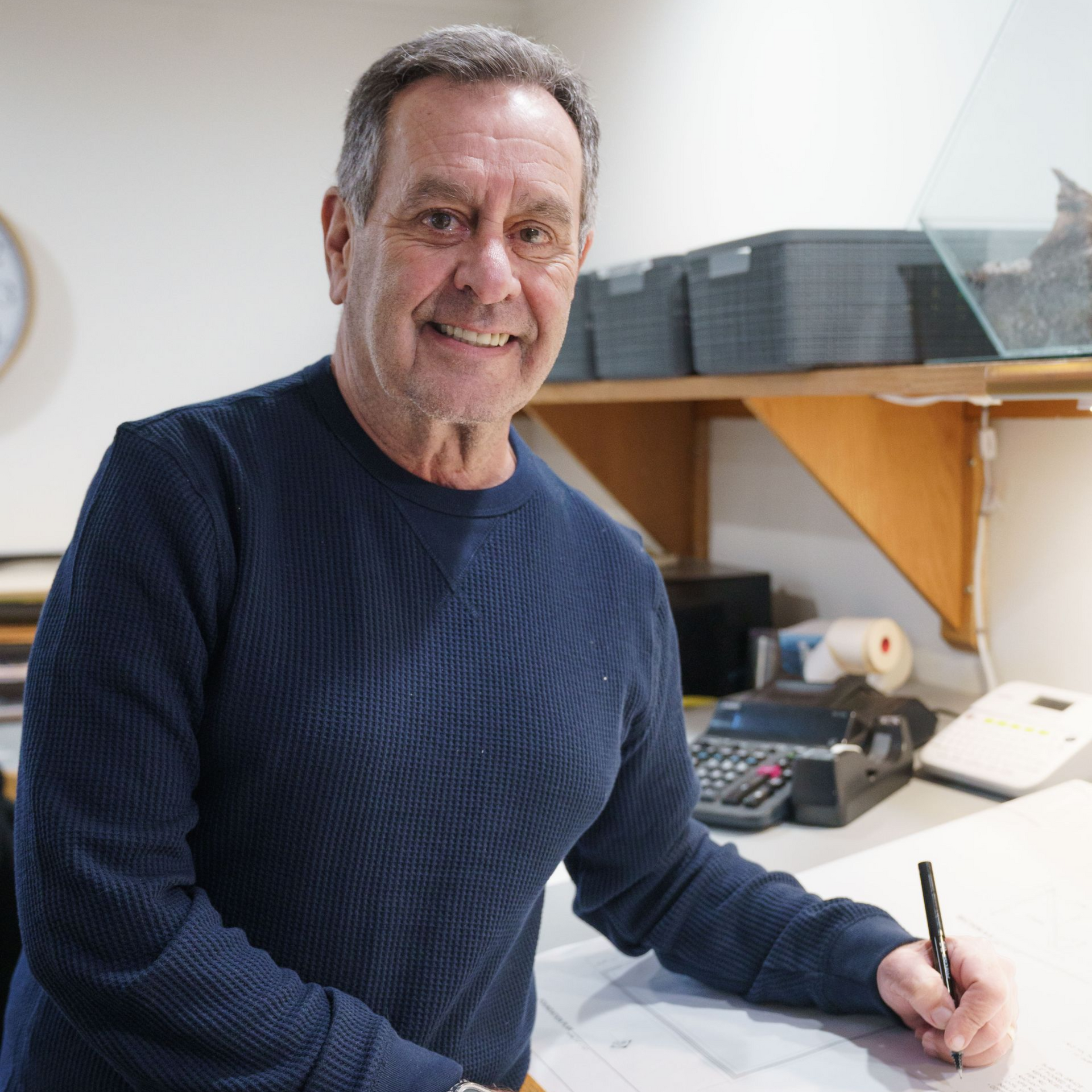 A smiling man wearing a blue sweater writes on a desk in an office with storage bins and office equipment in the background.