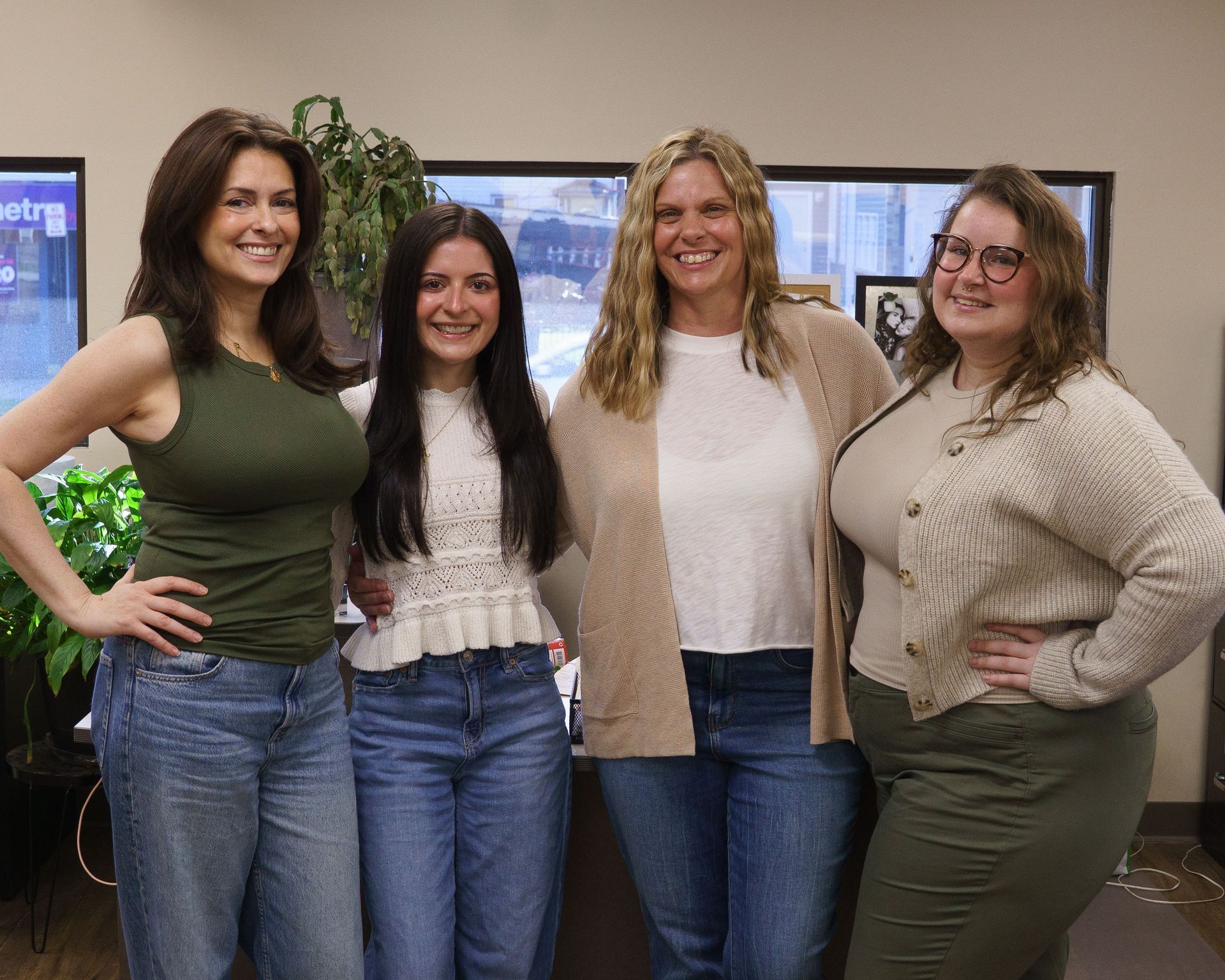 Four people pose smiling in an office, wearing casual clothing, standing and sitting near a desk.