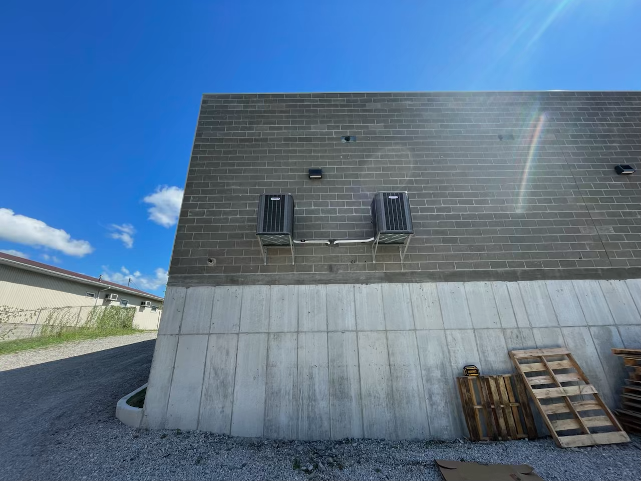 Two HVAC units mounted on a gray concrete block wall above a sloped concrete base, under a clear blue sky.