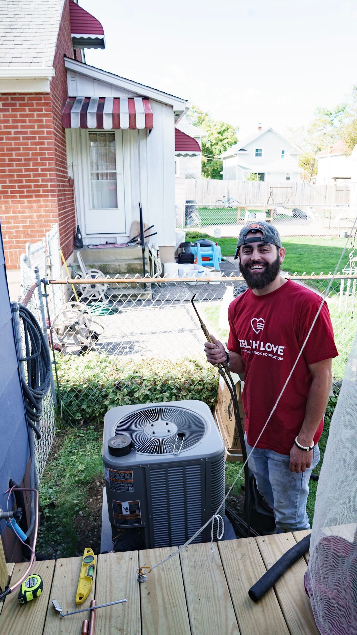 A smiling person in a red t-shirt stands outdoors next to an HVAC unit, holding a tool in their hand.