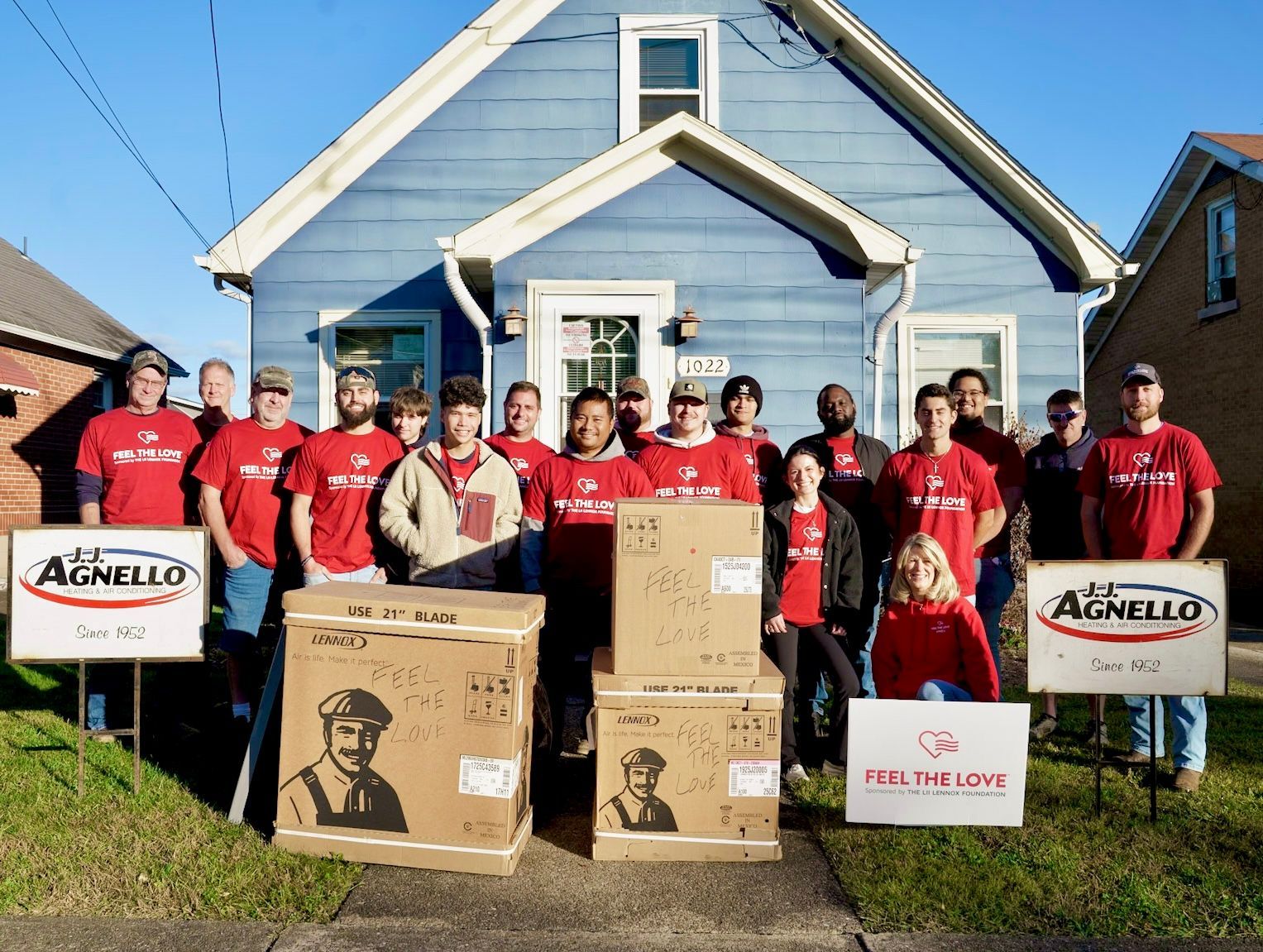 A large group poses in red shirts in front of a blue house with boxes,
