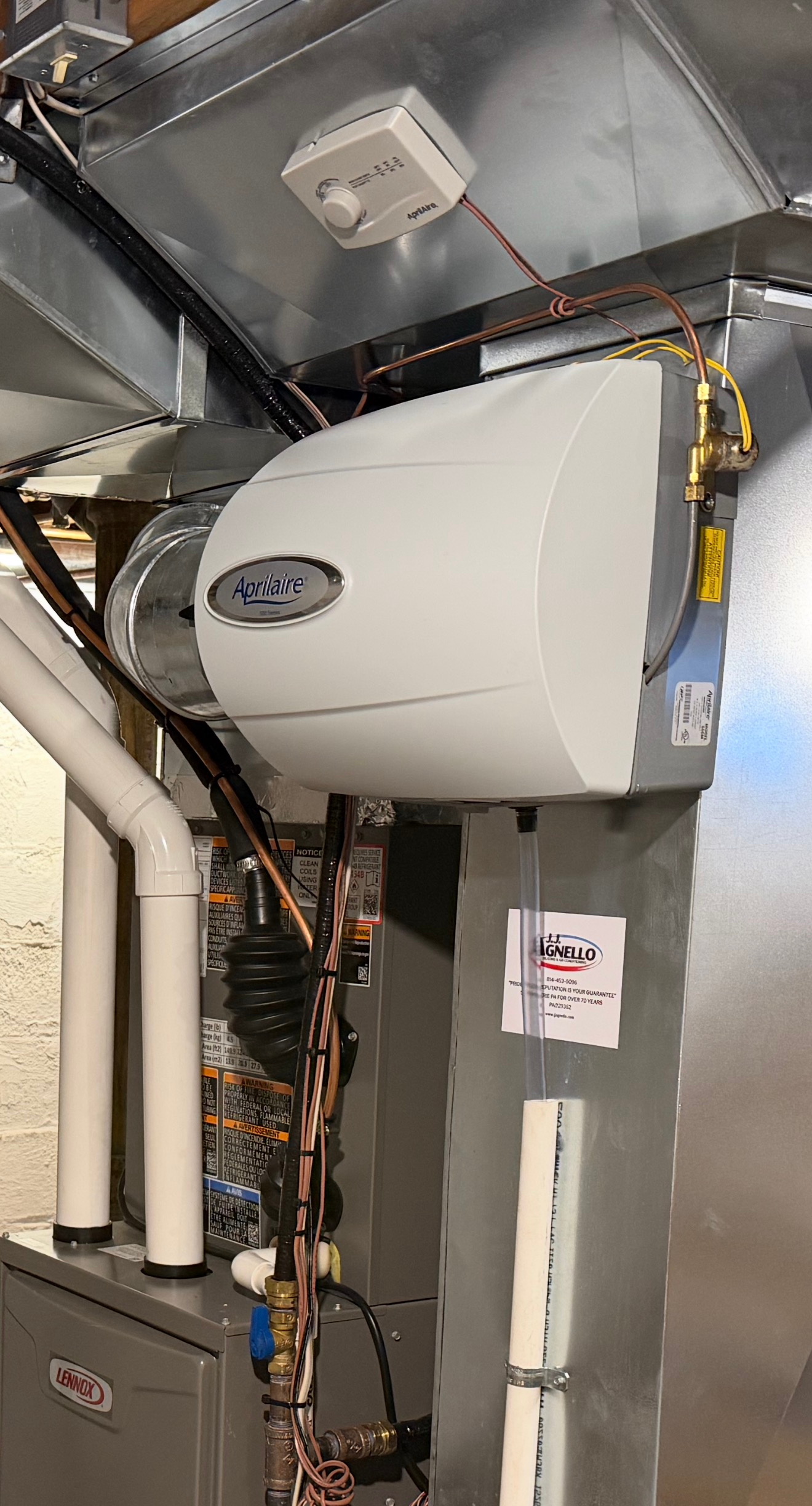 A white Aprilaire humidifier unit mounted to metal furnace ductwork in a basement setting.