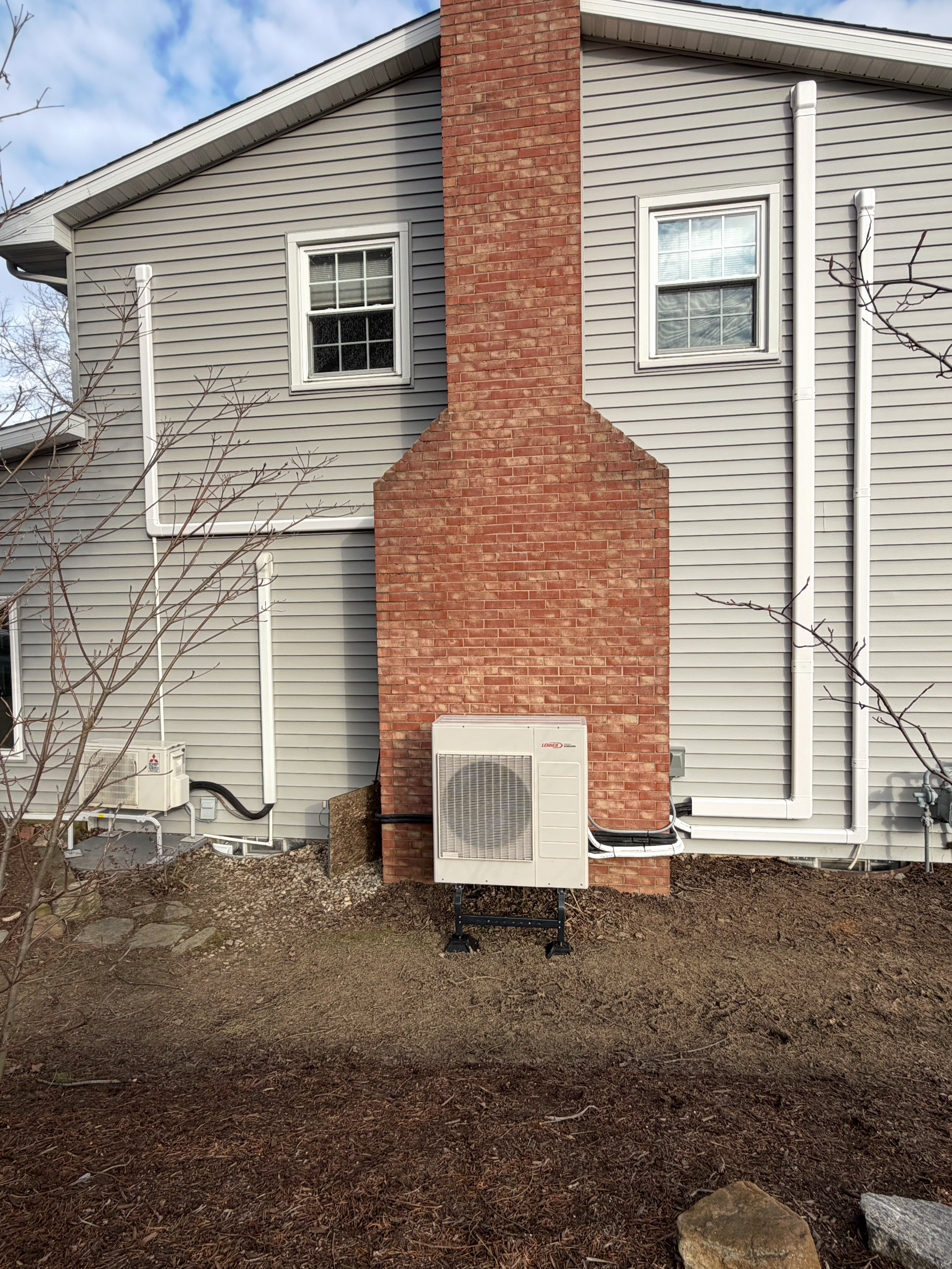 Exterior view of a light gray vinyl-sided house with a brick chimney and two HVAC units on a mulched ground.