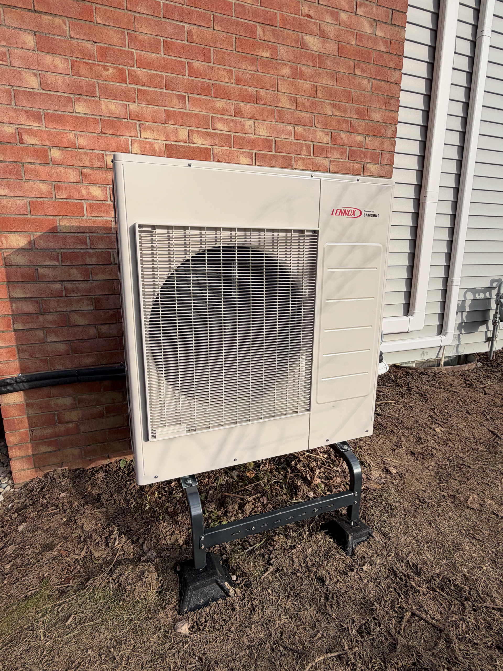 An outdoor HVAC condenser unit mounted on a black metal stand next to a brick wall and white siding.