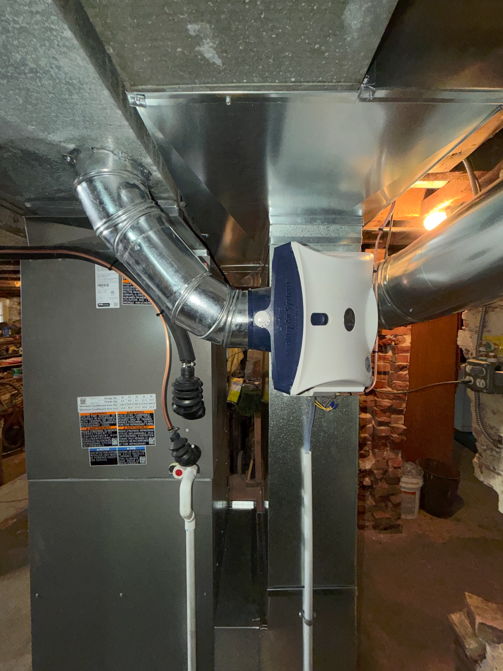 A white and blue humidifier unit mounted to a silver metal HVAC duct in a residential basement.