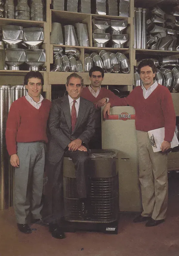 Four men stand in a metal ductwork warehouse, posing next to a heater. One man in a suit, three in red sweaters.