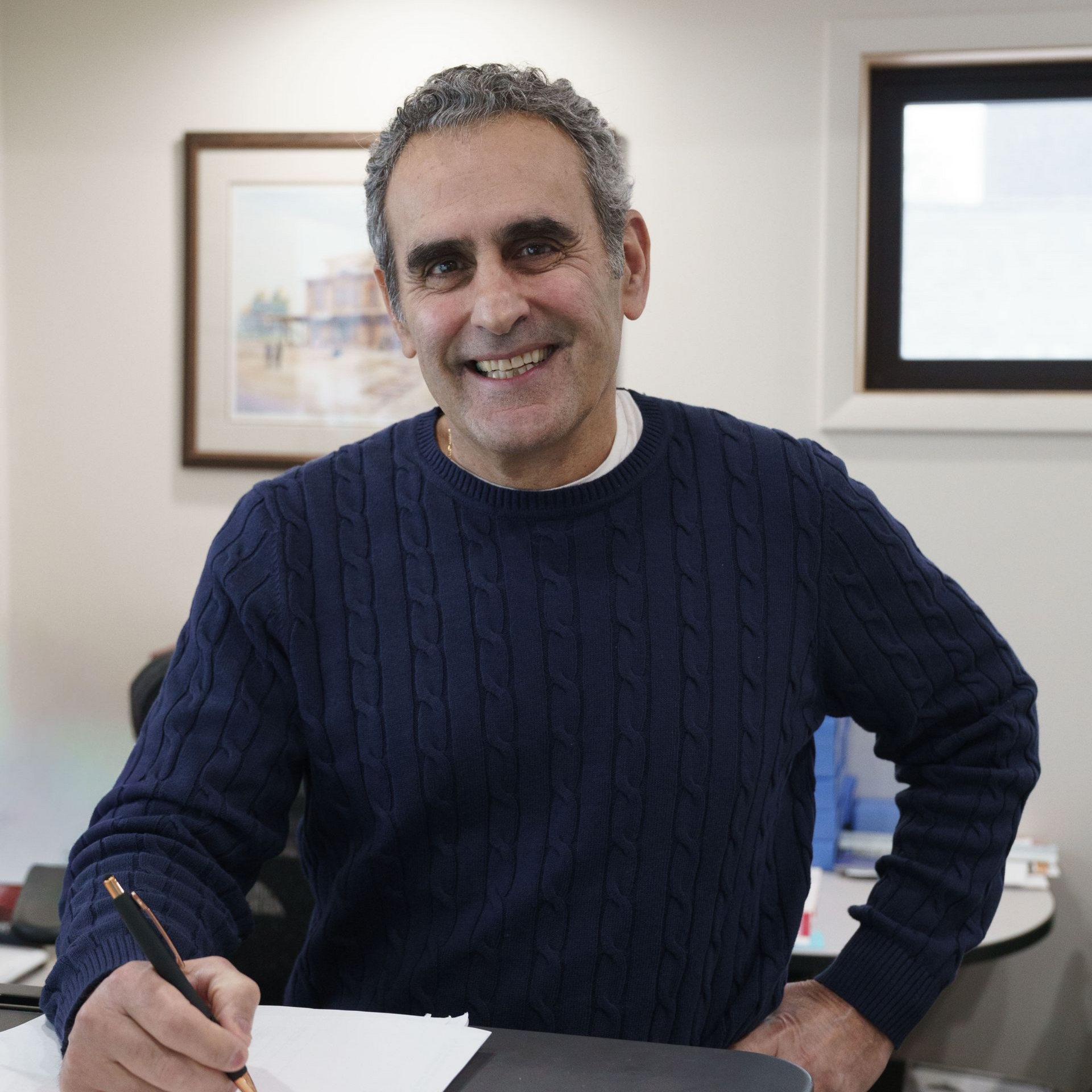 A person in a dark blue cable-knit sweater smiles while writing on a paper at a desk in an office.