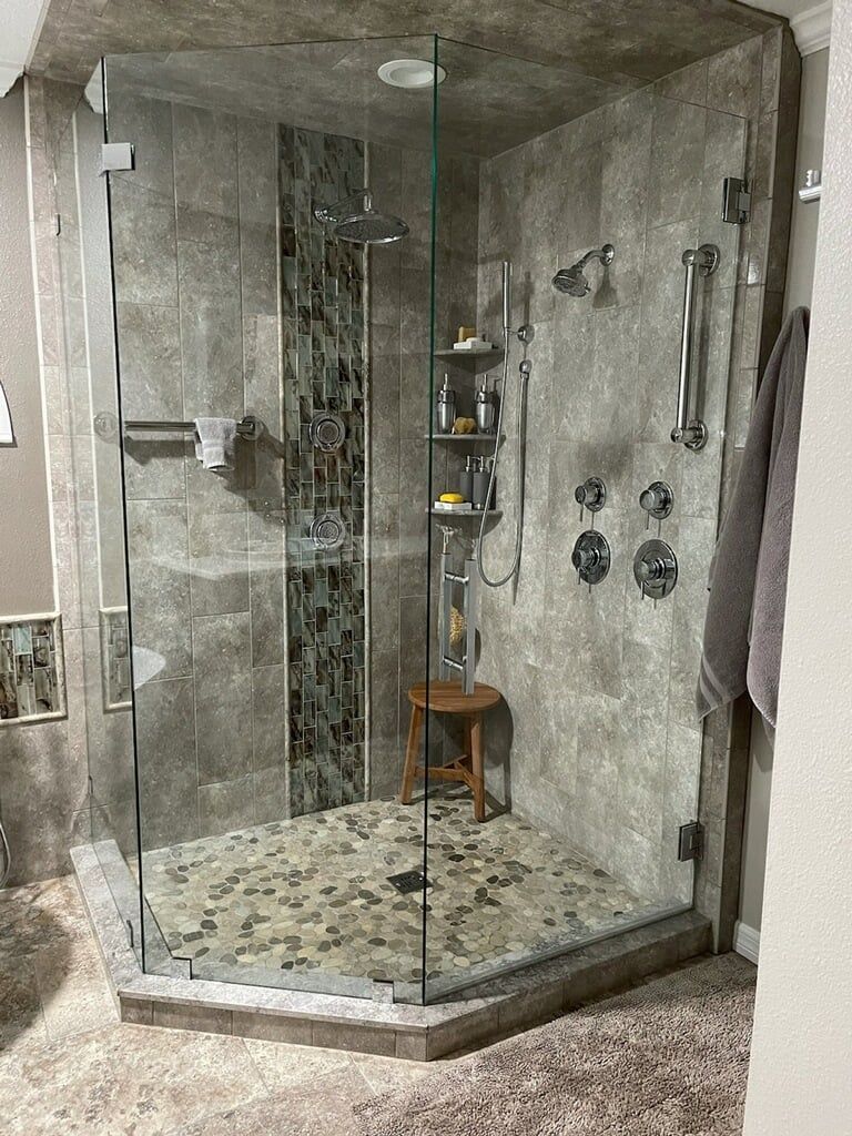 Corner glass shower stall with stone tile walls, pebble floor, and built-in shelves.