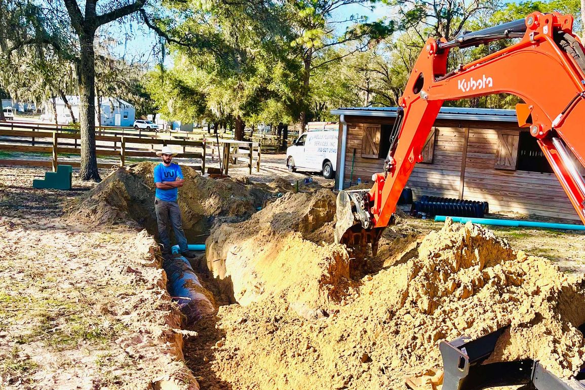 Excavator digging a trench beside a house, with a worker nearby in a wooded yard.