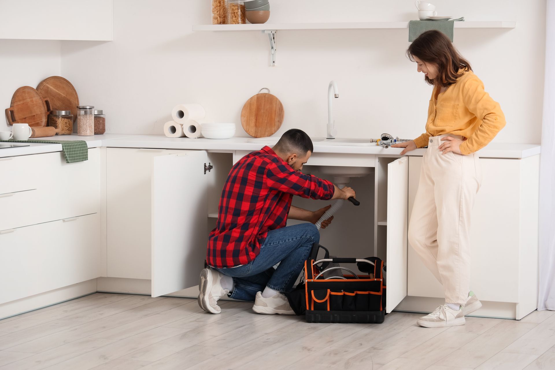 Person kneeling under kitchen sink, repairing plumbing while another stands nearby, in a bright white kitchen