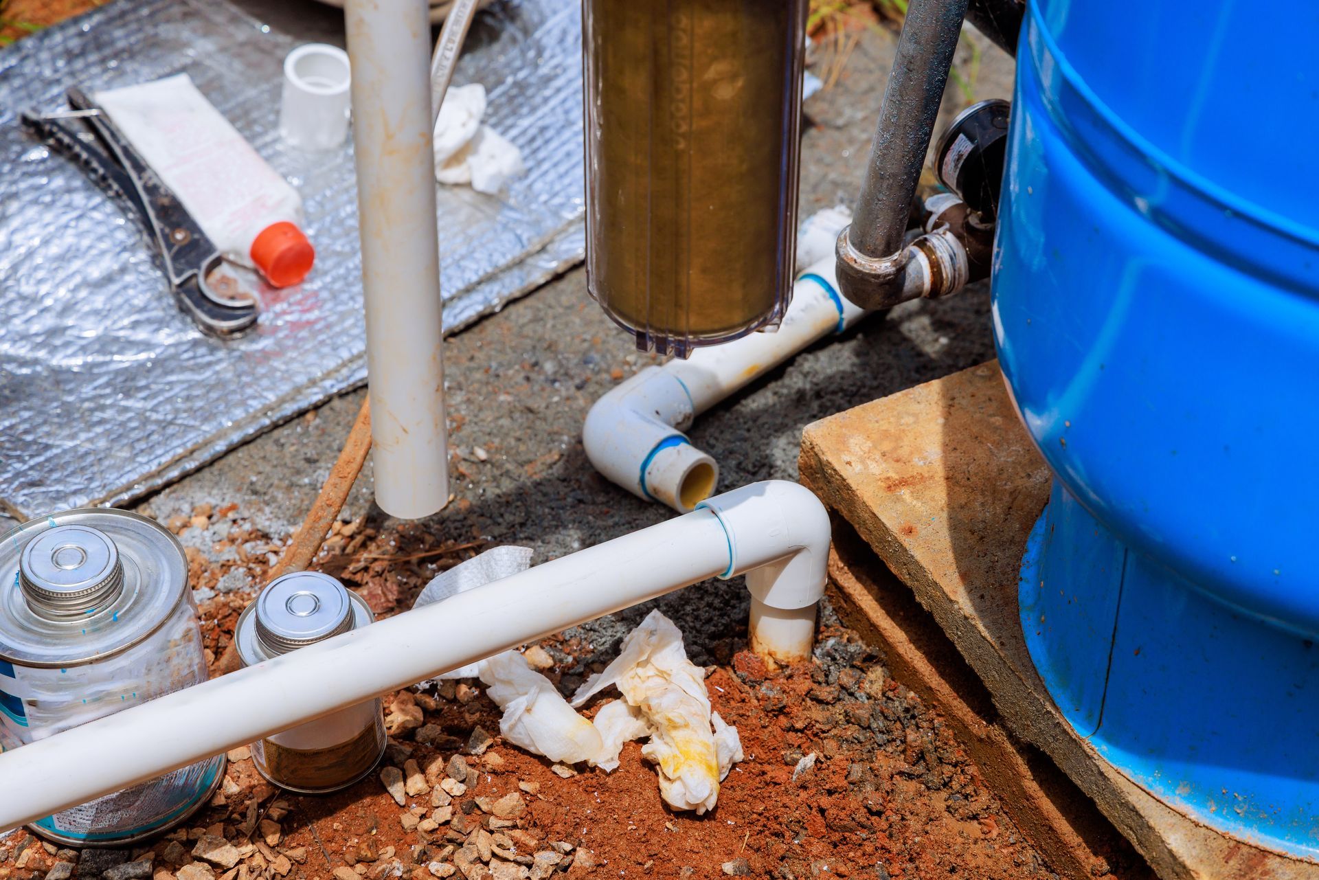 Plumbing pipes and valves beside a blue water tank on a rough outdoor floor